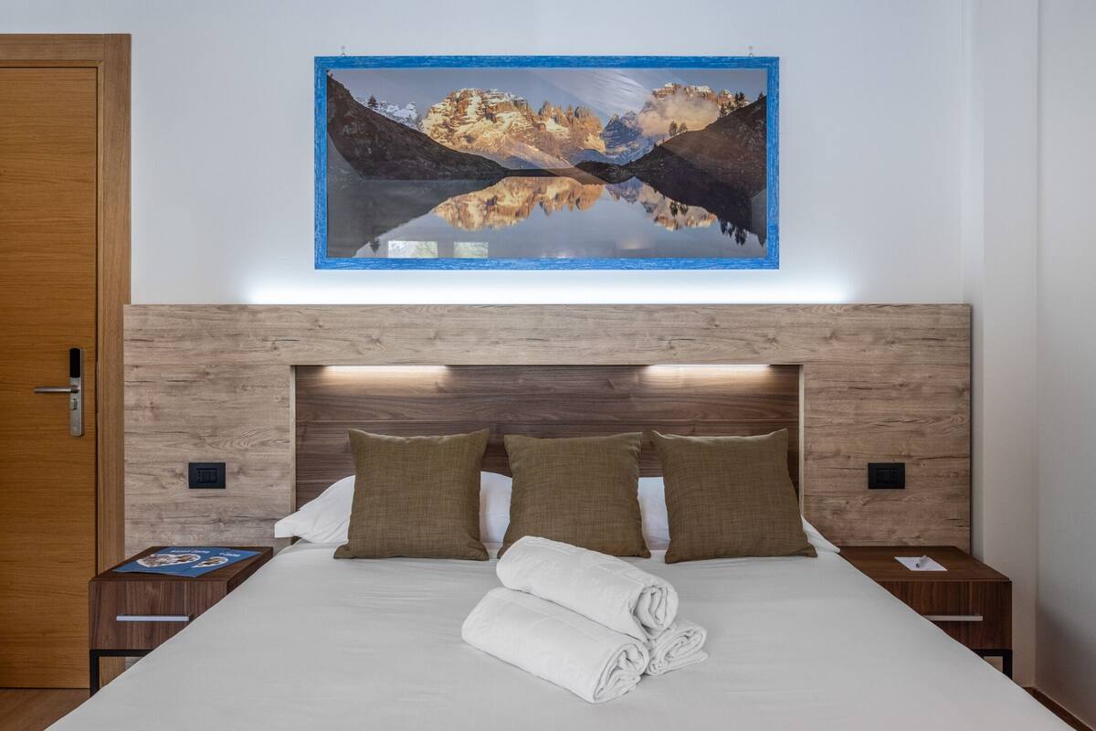Una cama cuidadosamente tendida con cabecera de madera, un cuadro colgado en la pared de un paisaje montañoso, y una mesita de noche de madera con un libro y un control remoto.