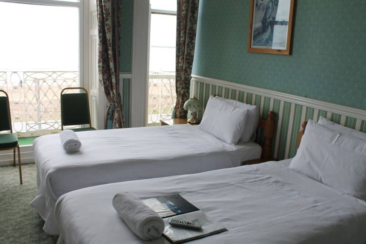 A hotel room with two beds, a window with curtains, a painting on the wall, and a chair by the window.