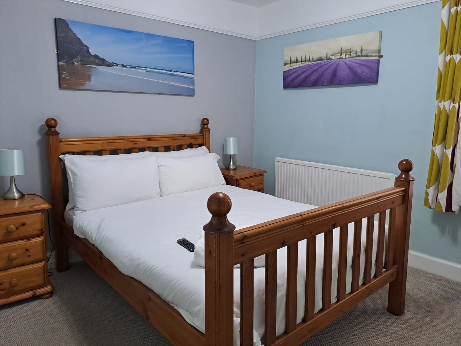 A bedroom with a wooden bed, two bedside tables with lamps, a radiator, and two wall-mounted paintings.