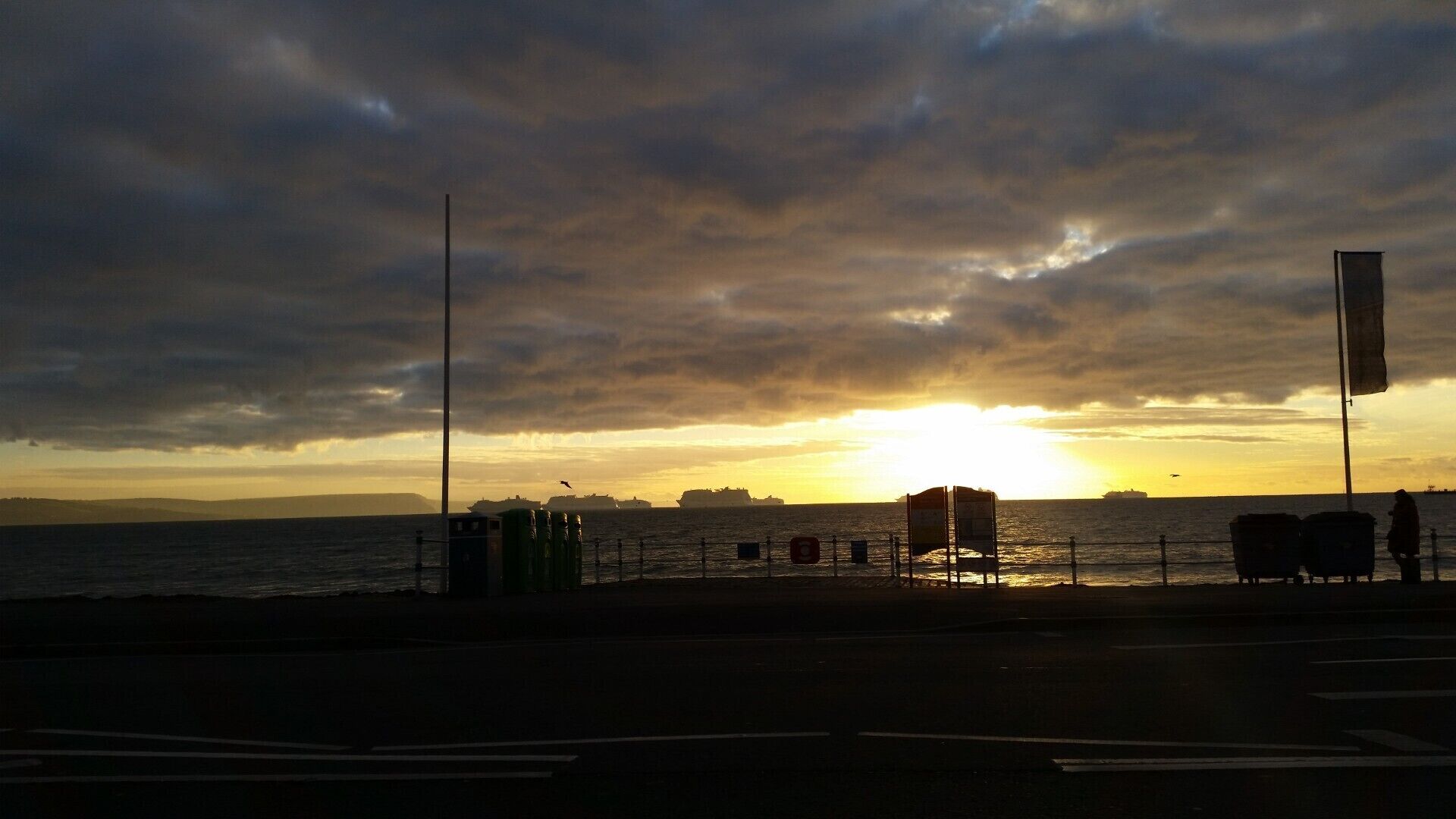 Un coucher de soleil sur une étendue d’eau, avec des navires visibles au loin, une clôture et un mât de drapeau.