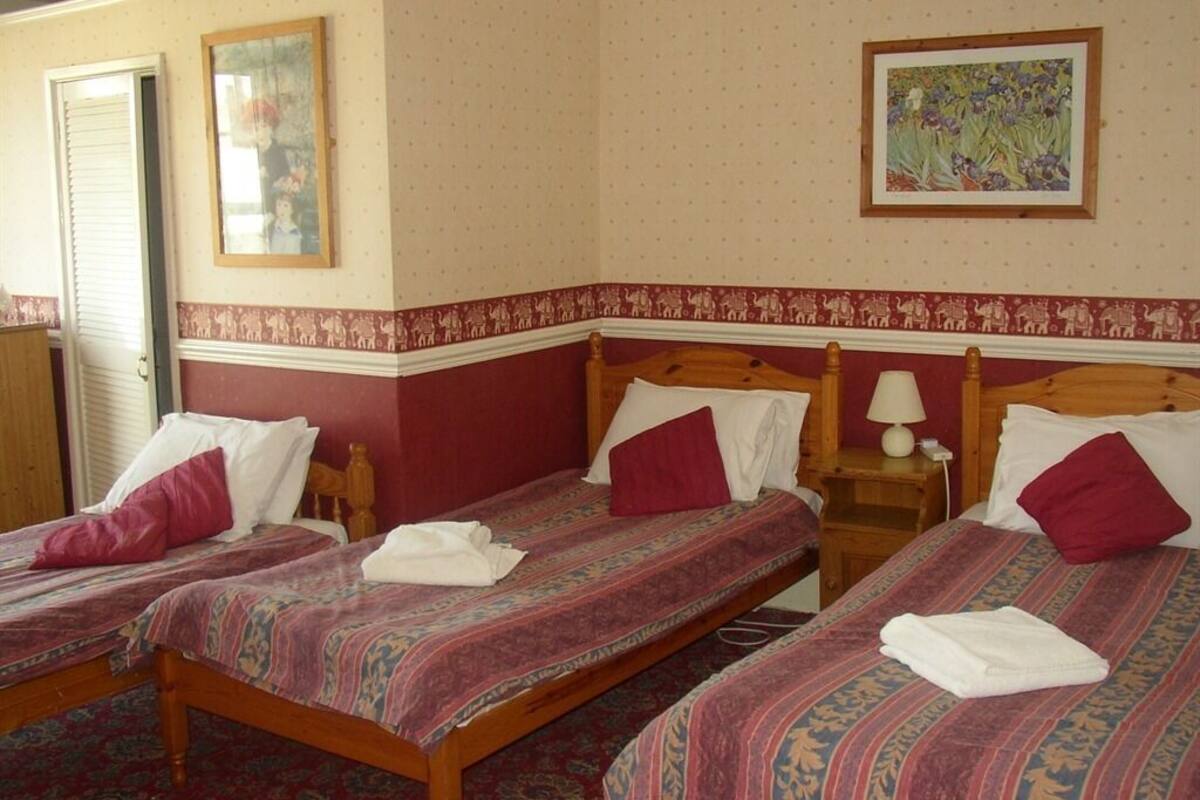 A hotel room with three wooden beds, each with a patterned bedspread and pillows. There is a nightstand with a lamp on one side of the room.