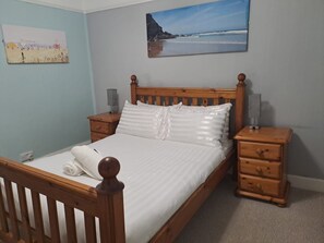 Une chambre à coucher avec un lit en bois, deux tables de chevet, une petite table d’appoint, une lampe fixée au mur et un tableau sur le thème de la plage.