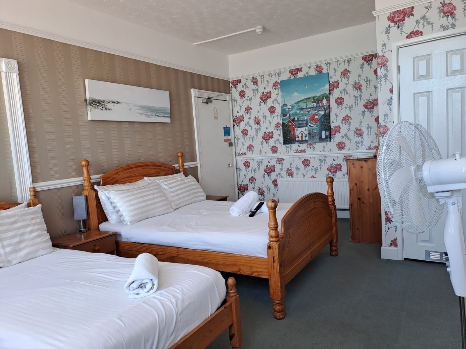 A hotel room with two wooden beds, a white fan, a painting on the wall, and floral wallpaper.