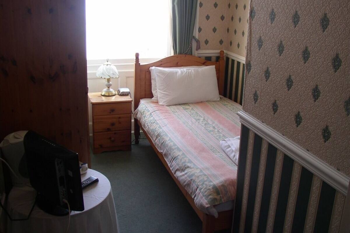 A small, cozy room with a single bed, a nightstand, a lamp, and a desk with a computer.