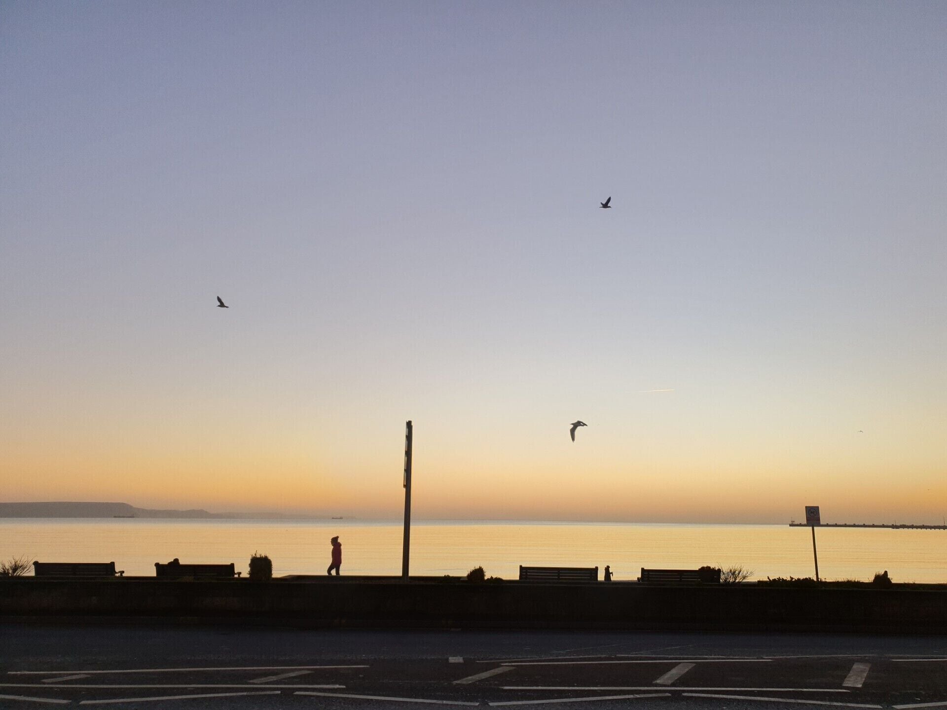 Une vue du coucher de soleil depuis un pont, avec une étendue d’eau calme, une personne qui se promène et des oiseaux qui volent.