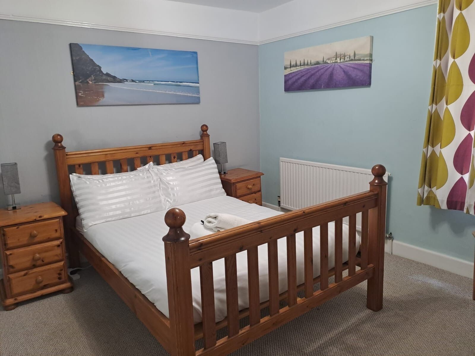 Un dormitorio con una cama de madera, dos mesitas de noche, un radiador y dos cuadros colgados en la pared.