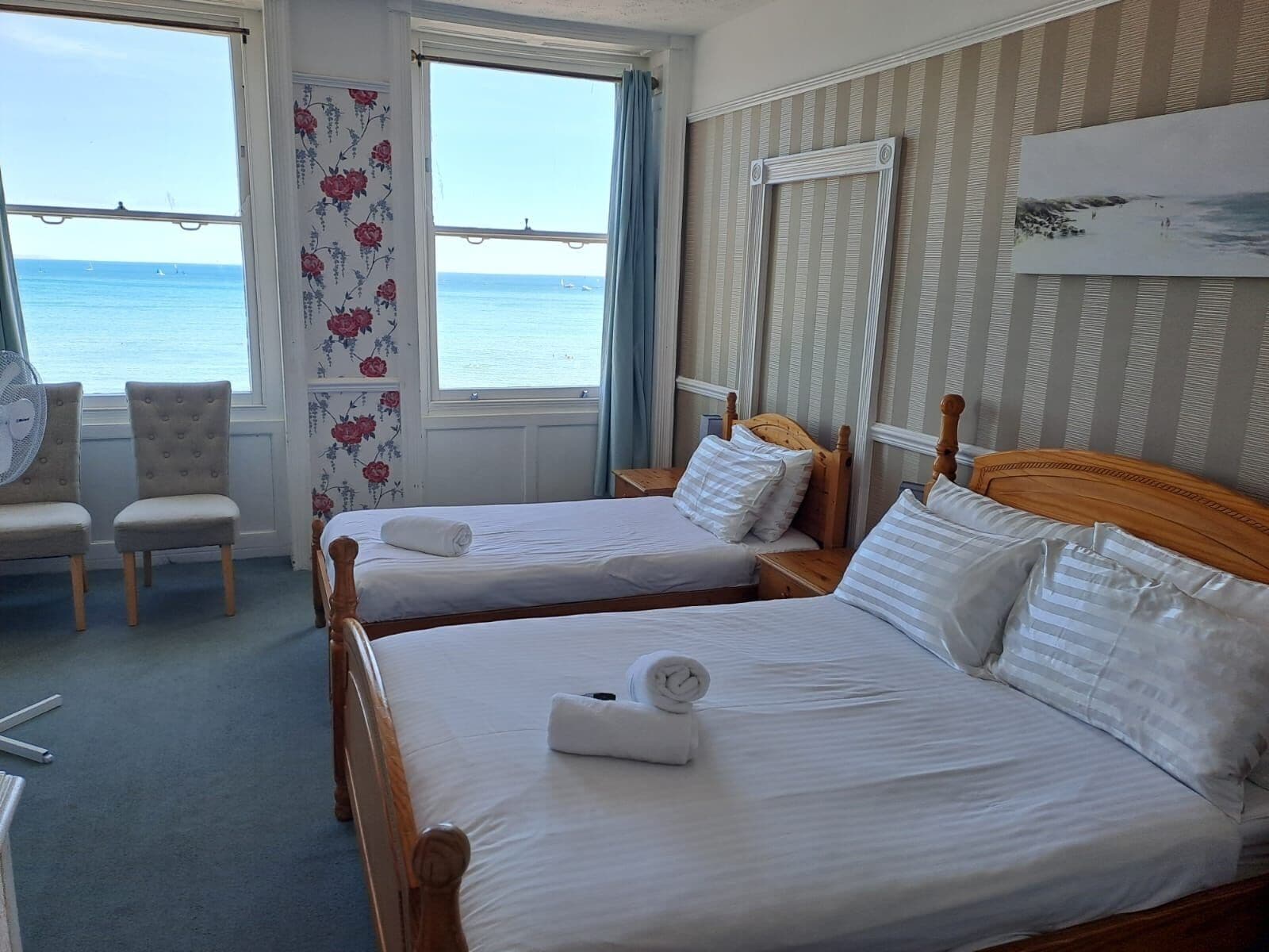 A hotel room with two beds, a chair, a fan, and a view of the sea.