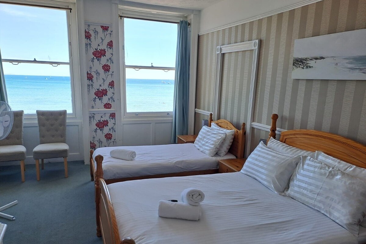 A hotel room with two beds, a chair, a fan, and a view of the sea.