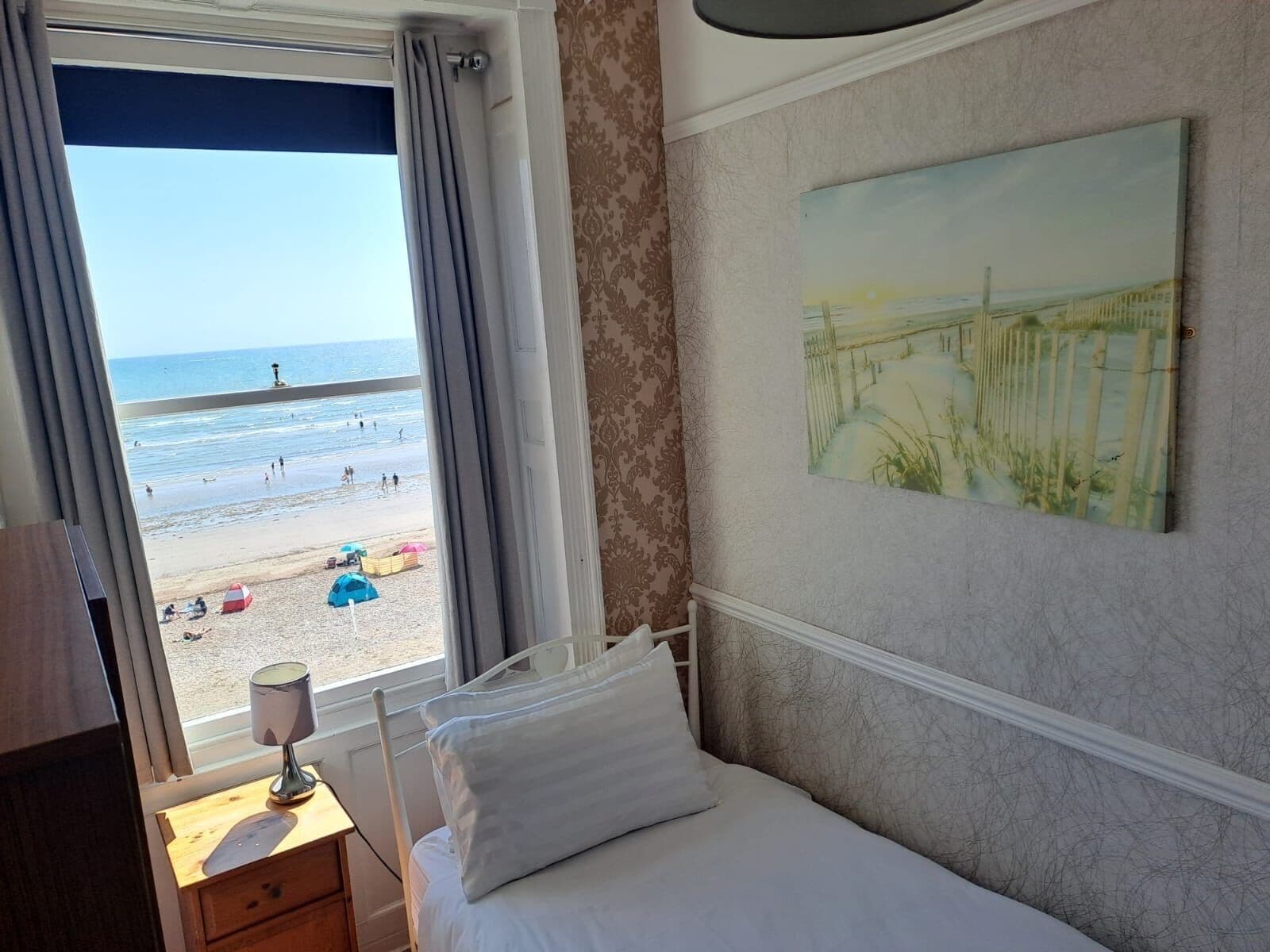Ein Hotelzimmer mit Bett, Nachttisch mit Lampe, einem Fenster mit Vorhängen und Blick auf den Strand.