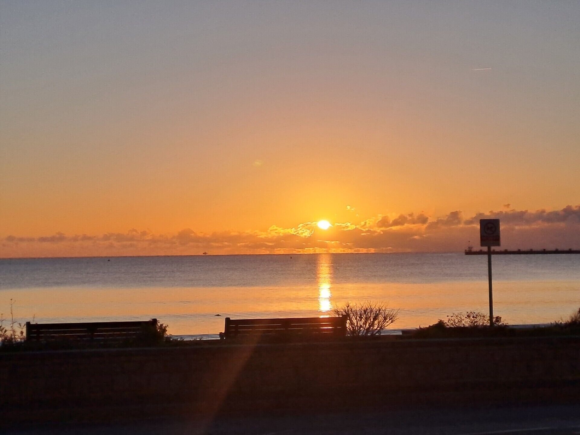 A sunset over a calm body of water with a bench and a signpost in the foreground.
