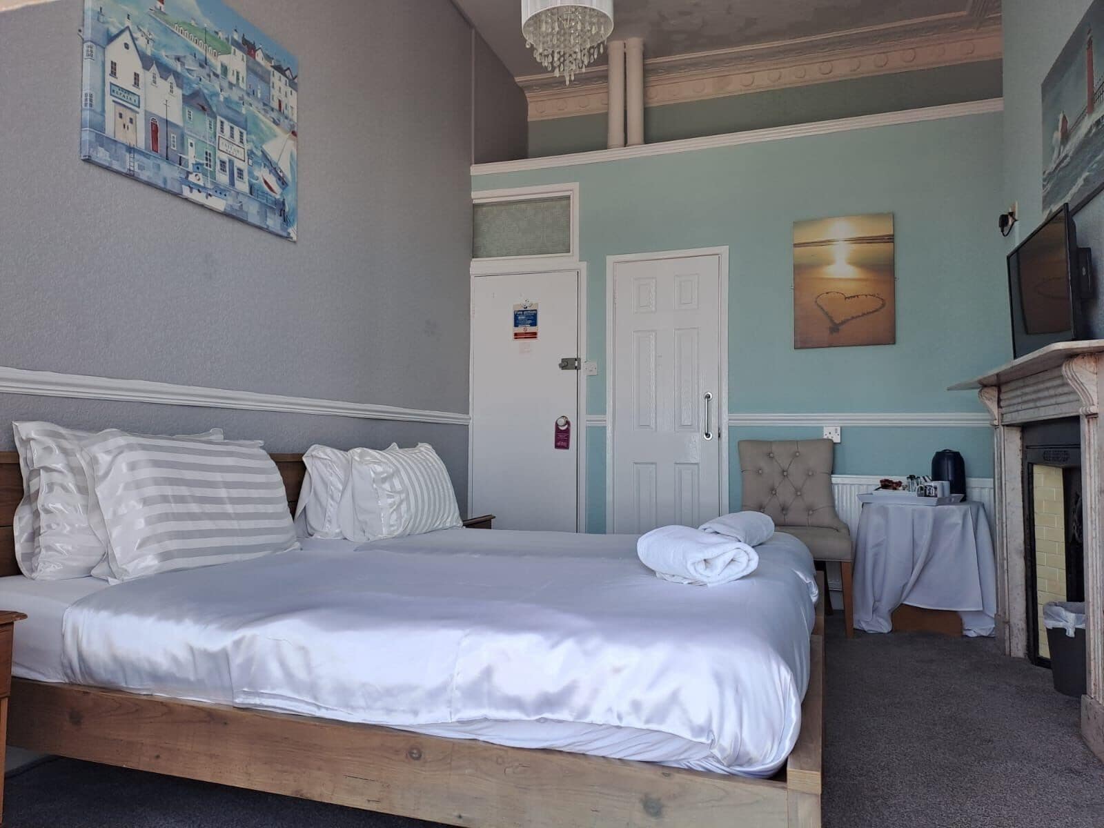 A hotel room with a bed, a painting, a fireplace, and a table with a kettle.