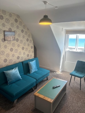 A living room with a blue sofa, a wooden coffee table, a blue armchair, a ceiling fan, and a window with a view of the sea.