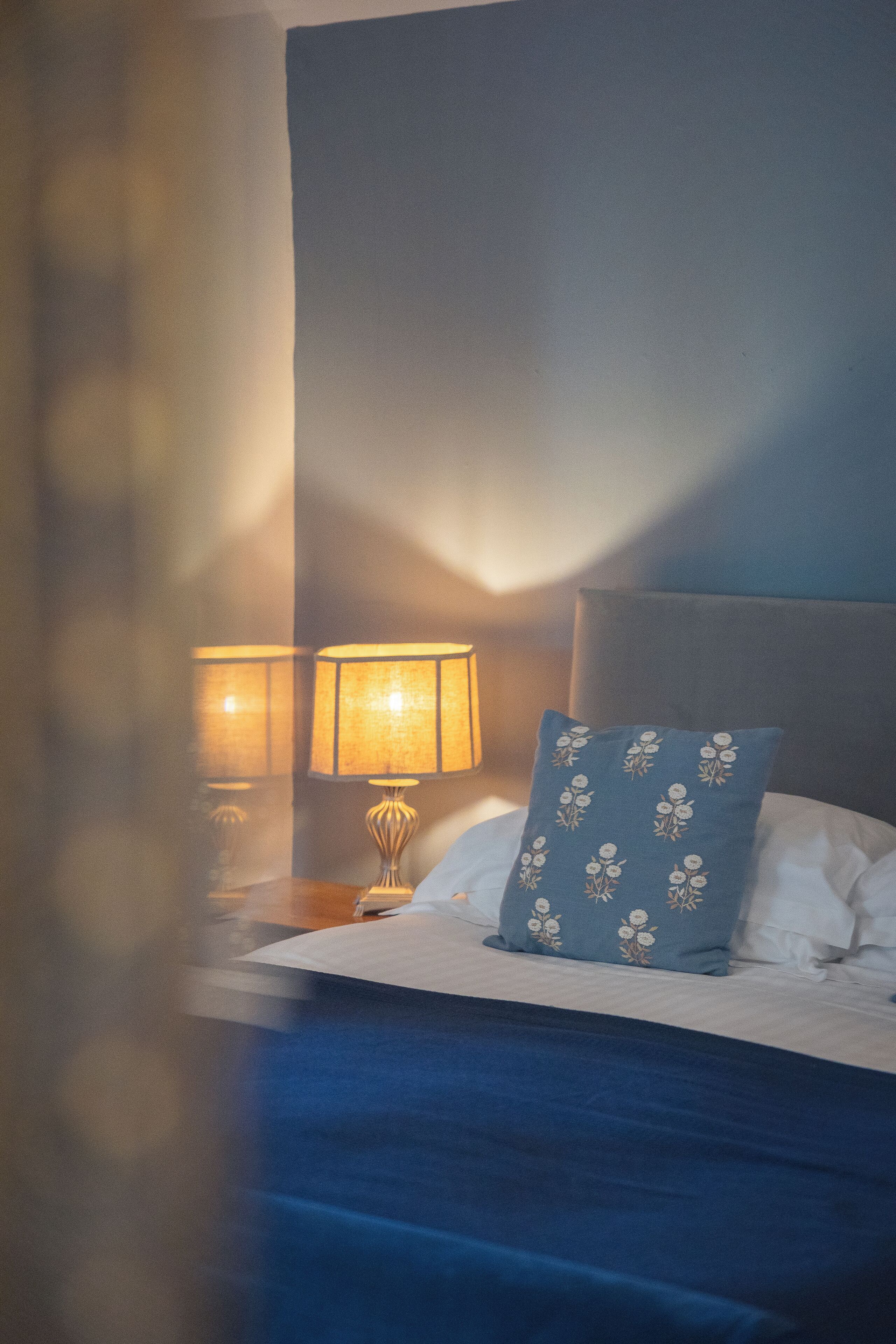 Una cama bien hecha con una colcha azul y una almohada estampada, una lámpara de noche y una mesita de noche.