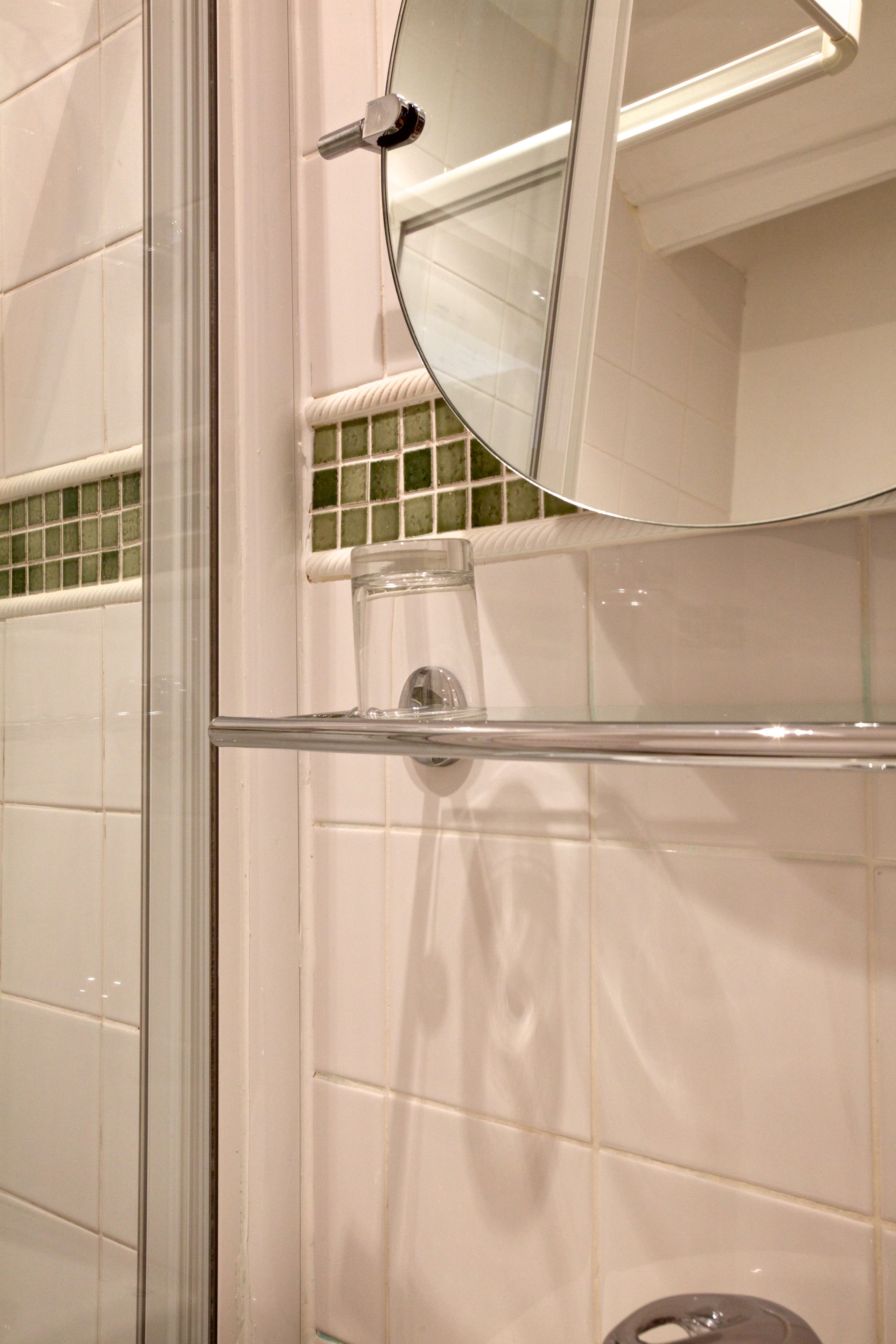 A bathroom with white tiles, a glass shower enclosure, a toilet with a glass tank, and a round mirror above the sink.