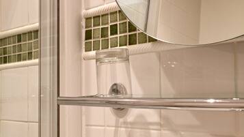 A bathroom with white tiles, a glass shower enclosure, a toilet with a glass tank, and a round mirror above the sink.