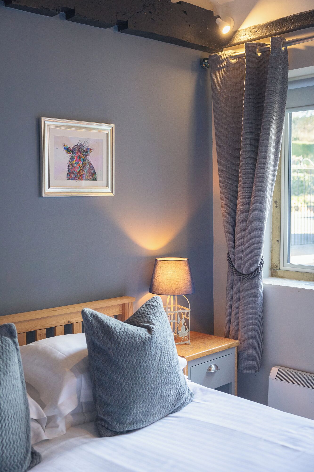 Een slaapkamer met een bed, kussens, een nachtkastje met een lamp, een lijst met een afbeelding aan de muur, en een raam met gordijnen.