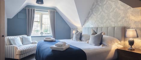 A bedroom with a large bed, a smaller sofa, a bedside table with a lamp, and a window with curtains.