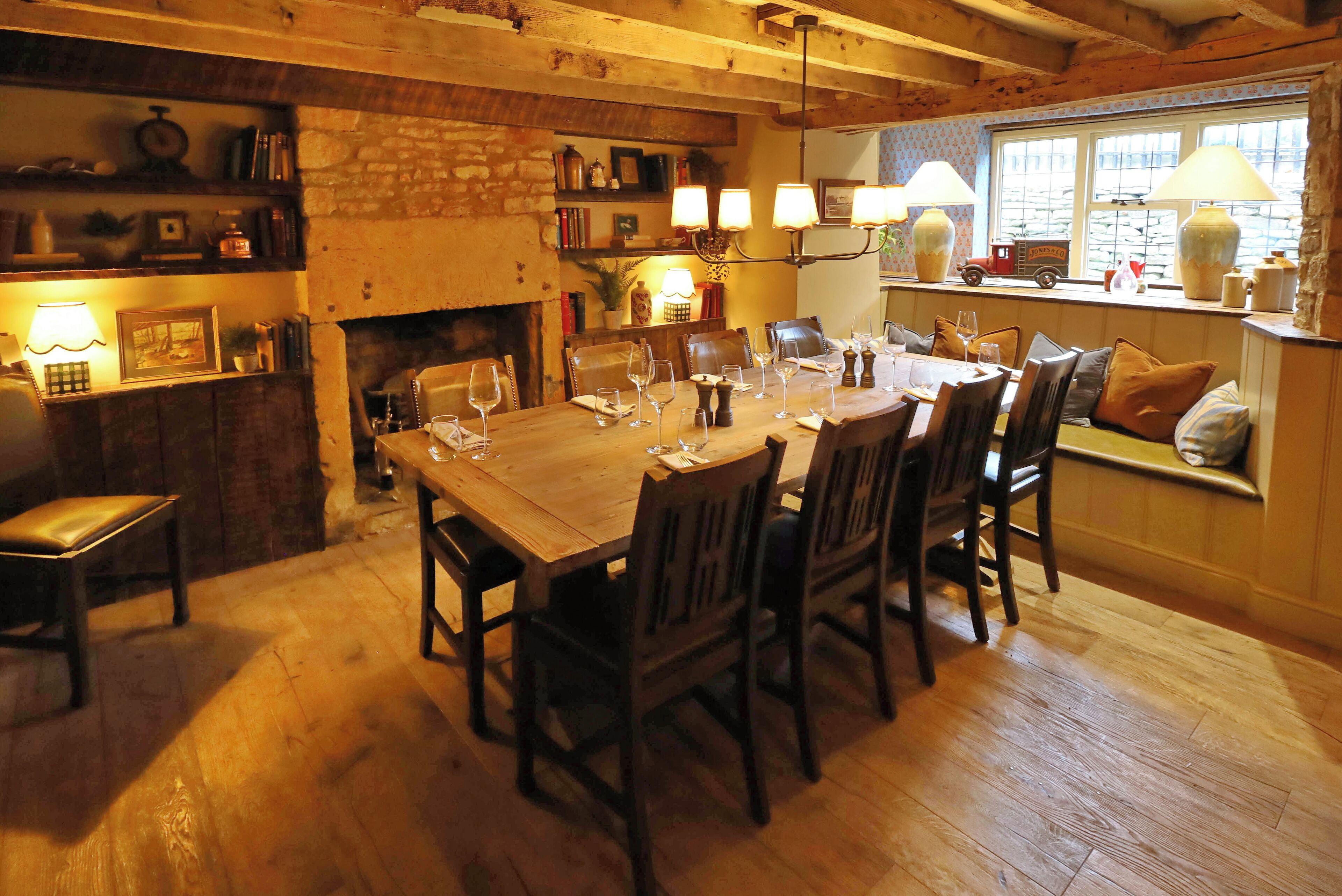 Un'accogliente sala da pranzo con un tavolo di legno apparecchiato per sei persone, circondato da sedie. Sullo sfondo si nota un camino e scaffali con diversi oggetti, e una finestra che offre vista sull'esterno.
