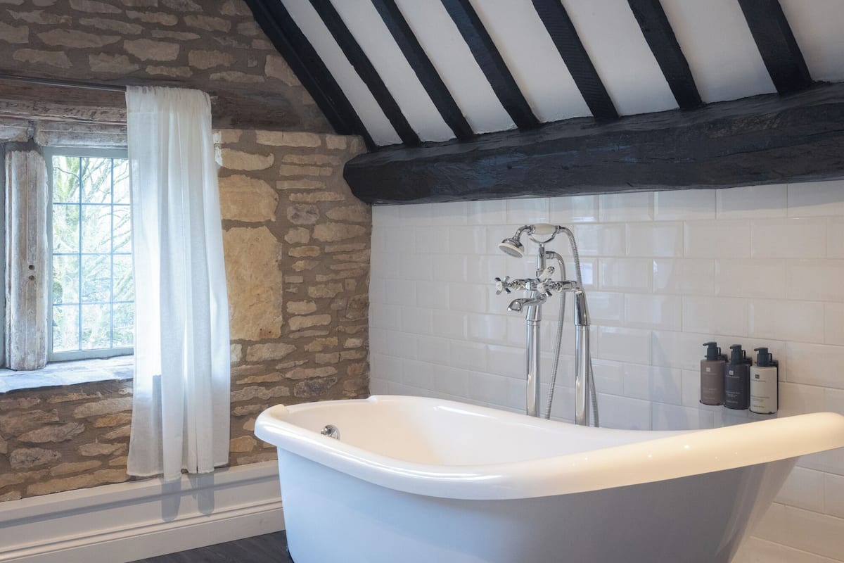 A bathroom with a freestanding bathtub, stone walls, a window with a view, and a black beam ceiling.