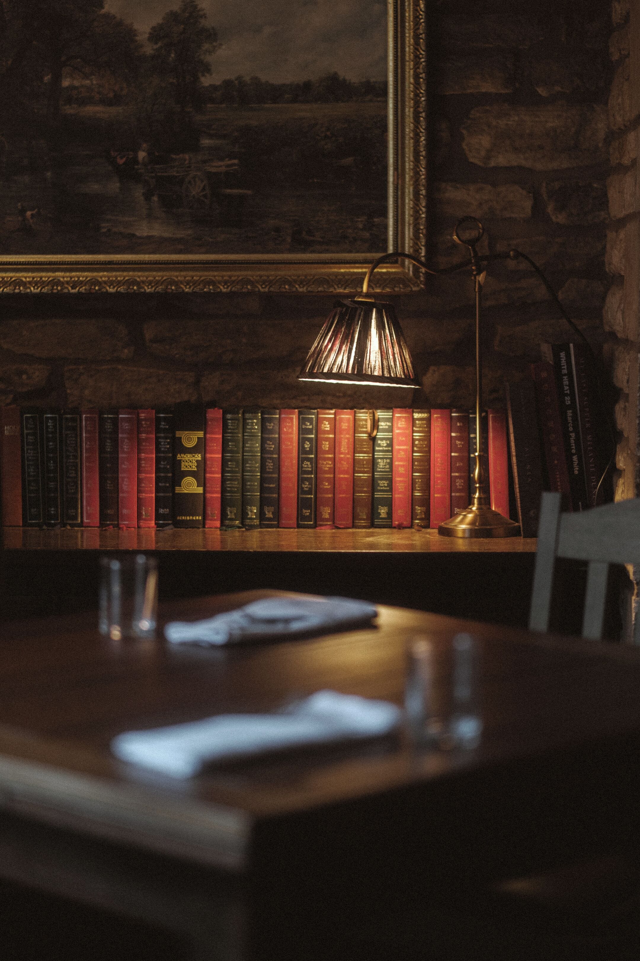A dimly lit room with a wooden table, a lamp, and a bookshelf filled with books.