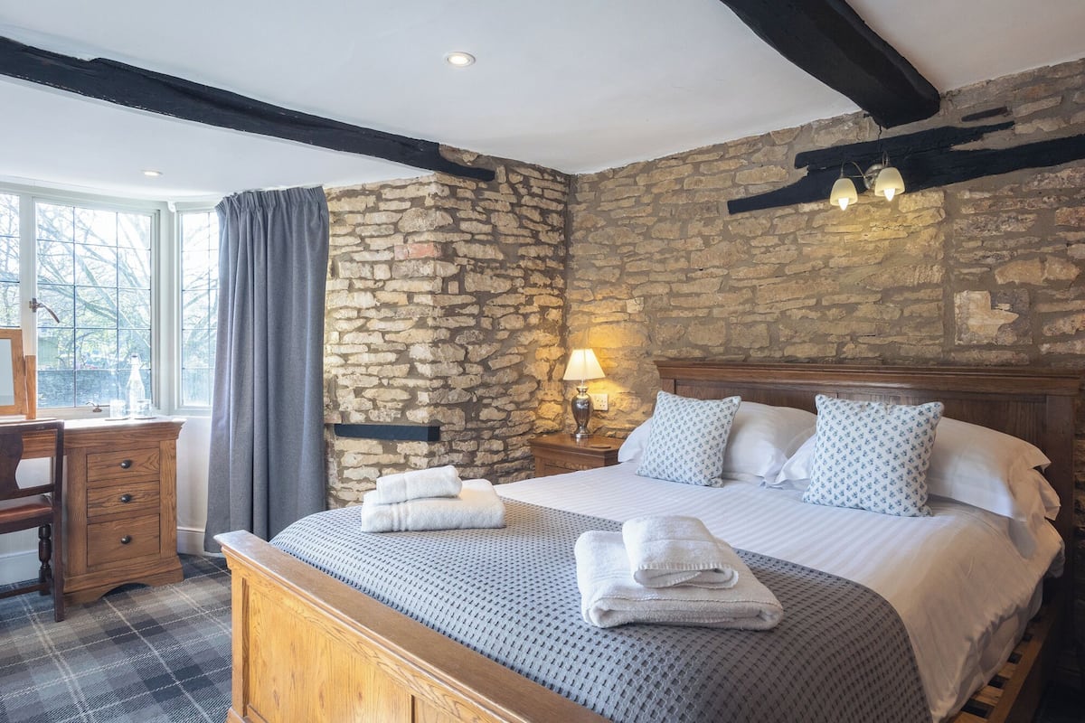 A bedroom with a stone wall, a wooden bed, a nightstand with a lamp, a desk with a chair, and a window with curtains.