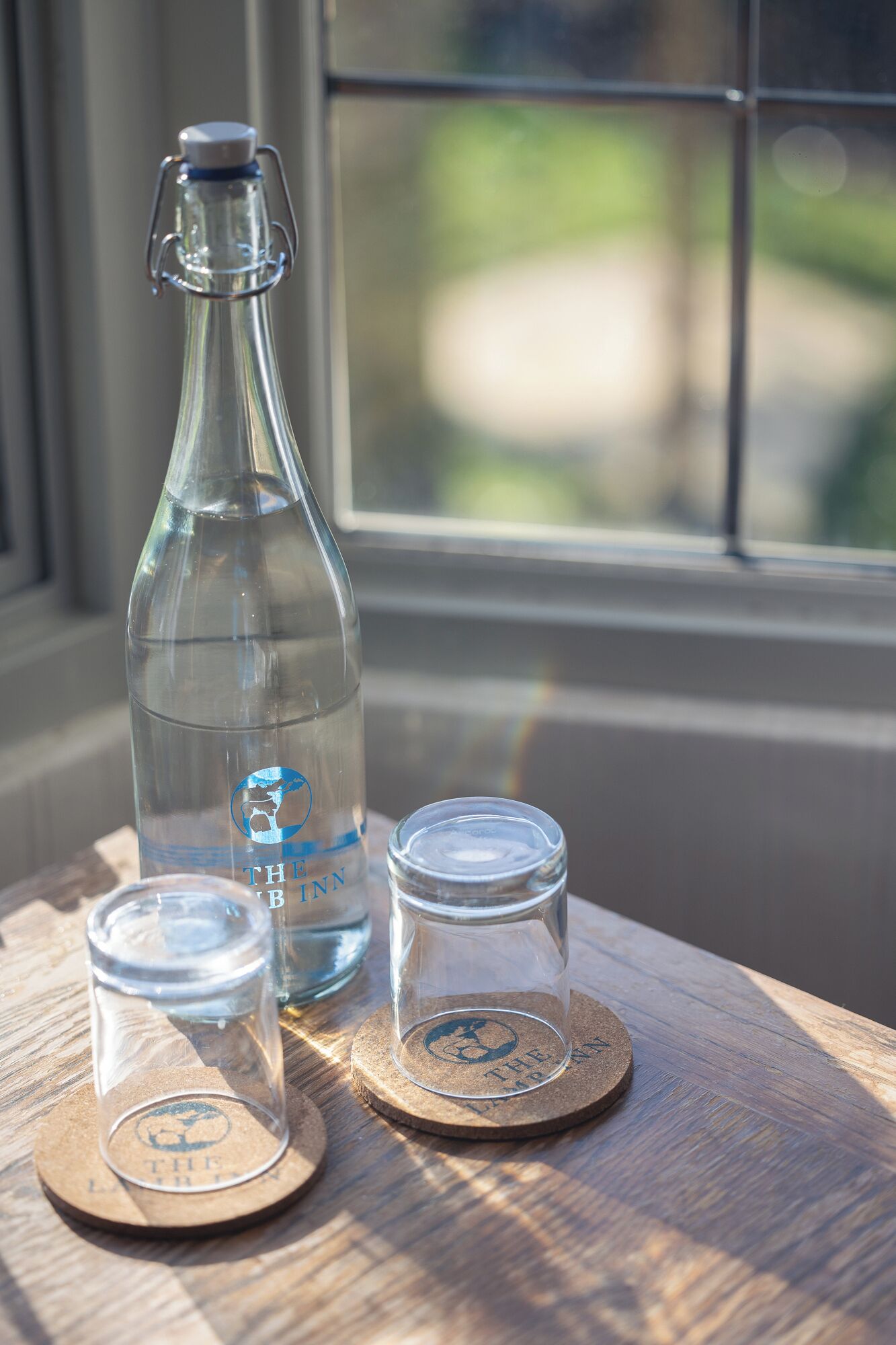 A clear glass bottle with a metal clasp and a metal lid, two small glass cups with lids, and two coasters with a logo on a wooden surface by a window.