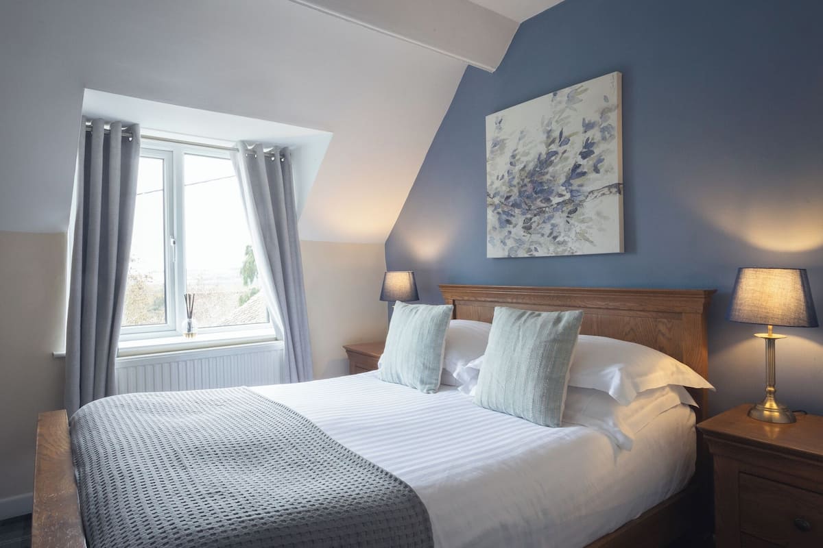 A bedroom with a large bed, two bedside tables, a window with curtains, a wall-mounted painting, and a lamp on the nightstand.