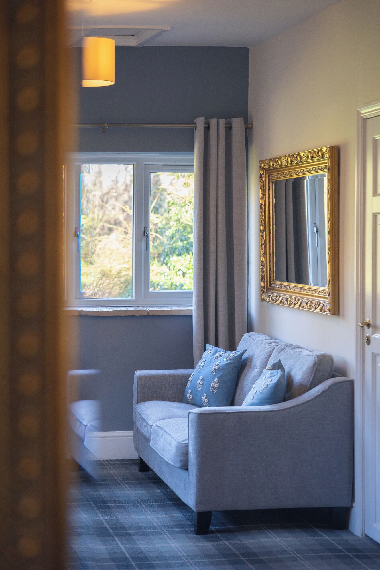 A room with a grey sofa, a large mirror with a gold frame, and a window with curtains.
