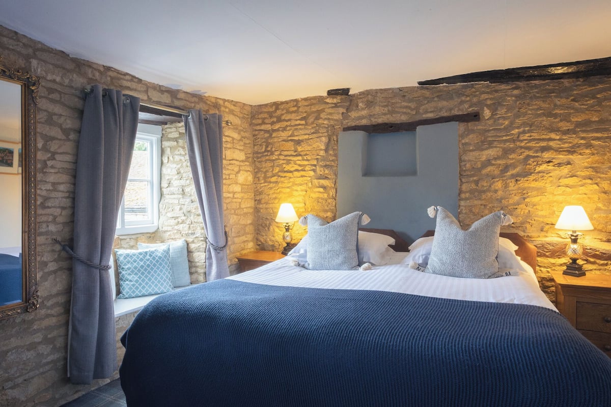 A bedroom with a stone wall, a bed with a blue blanket, two bedside lamps, and a window with curtains.