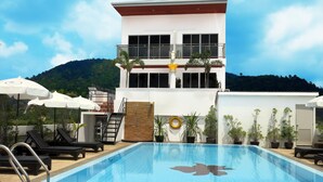 Una piscina con sillones de descanso y sombrillas, un edificio blanco con balcón, y montañas al fondo.