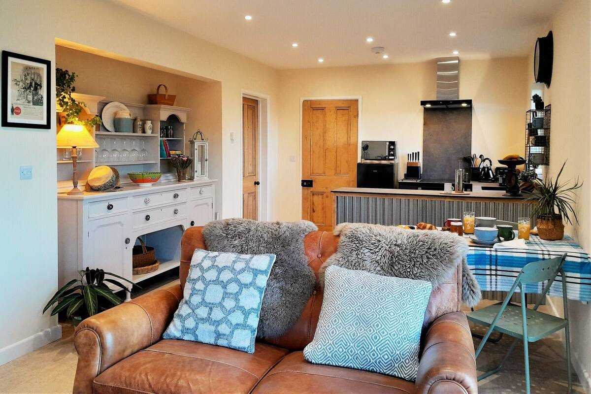 A cozy living room with a brown leather sofa, patterned cushions, and a dining area with a blue and white checkered tablecloth.