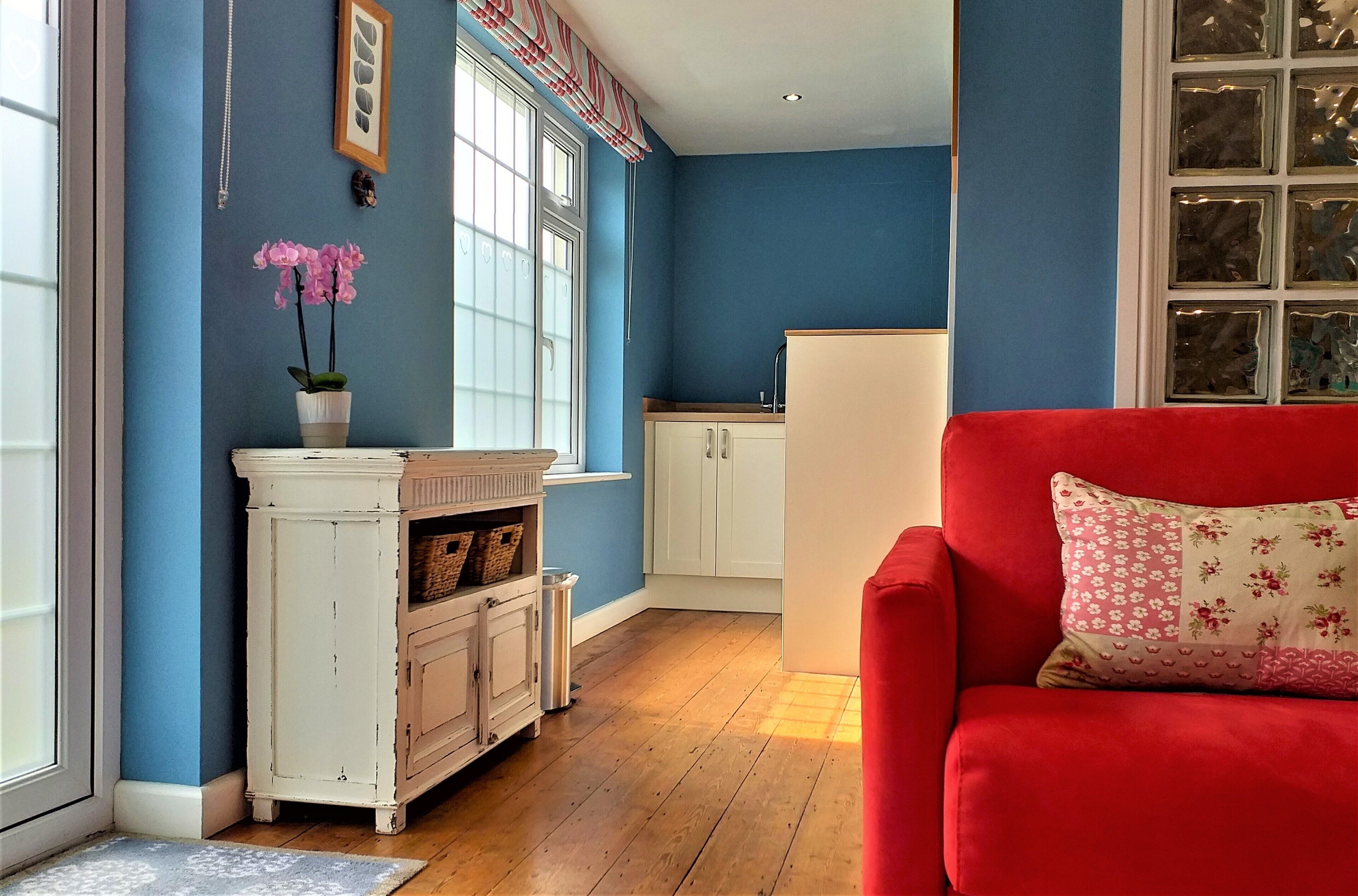 A room with a red sofa, a white cabinet with baskets, a window with curtains, and a kitchen area with cabinets.