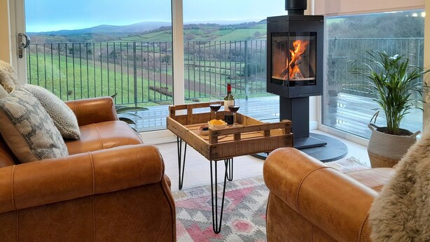 A cozy living room with leather sofas, a wooden coffee table with drinks, and a large window overlooking a scenic landscape. - Lobhill Farmhouse (Okehampton)