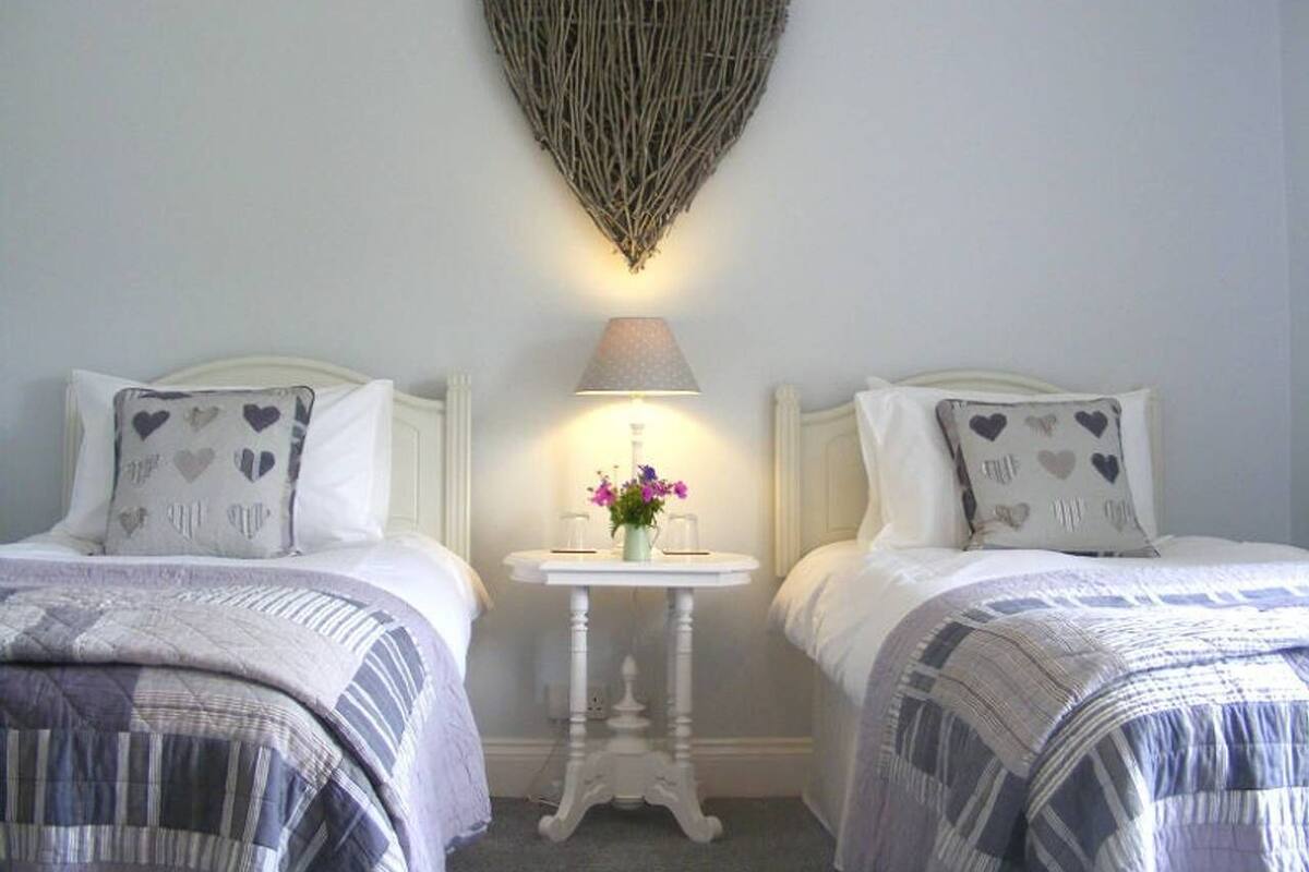 A bedroom with two beds, a small table with a lamp, and a heart-shaped decoration on the wall.