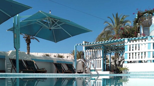 A clear blue pool with lounge chairs and umbrellas, surrounded by a white fence and palm trees. - Hotel Le Beau Sejour (Mezraia)