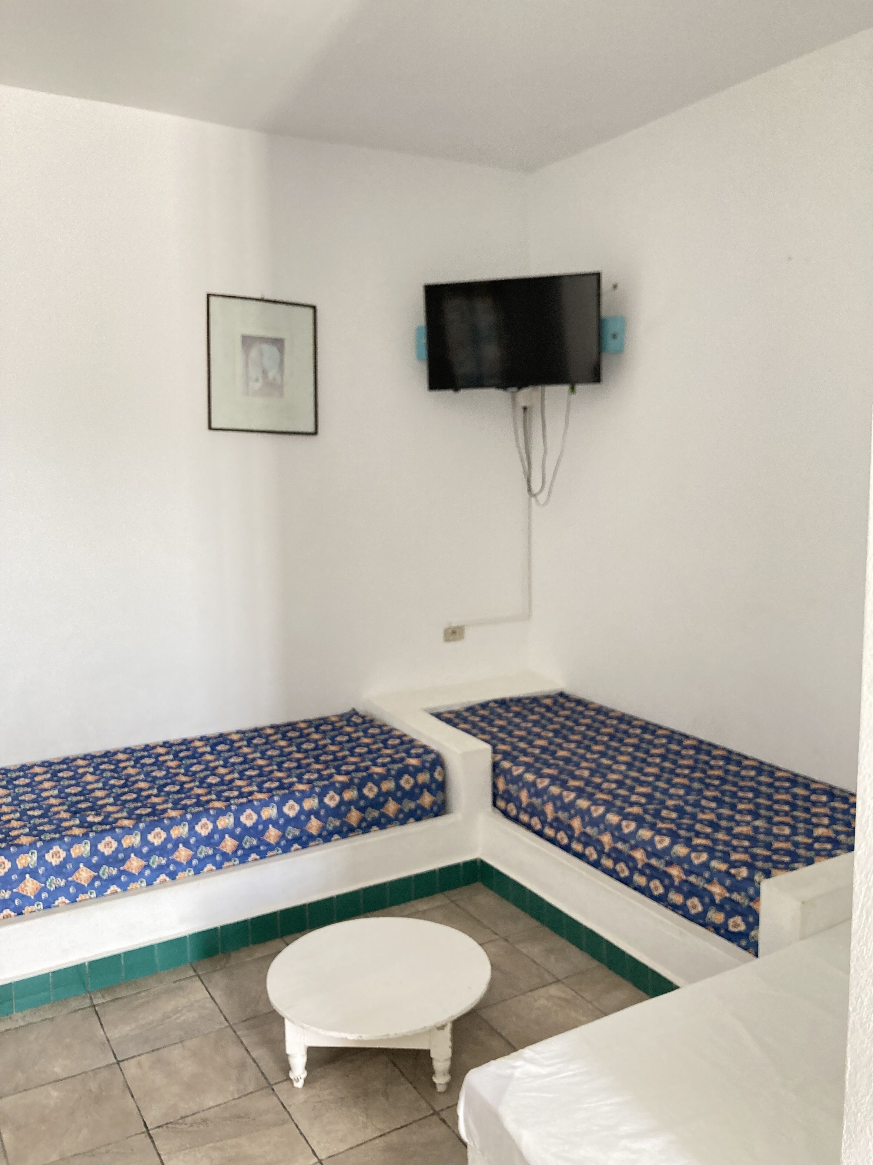 A small room with two single beds with patterned bedding, a white table, and a television mounted on the wall.