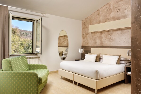 A bedroom with a large bed, a green armchair, a window with a view of trees, and a mirror on the wall. - Recostano (Trevignano Romano)