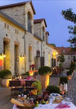 Un espace de restauration en plein air avec une table dressée pour un repas, entouré de bâtiments en pierre et de sièges extérieurs.