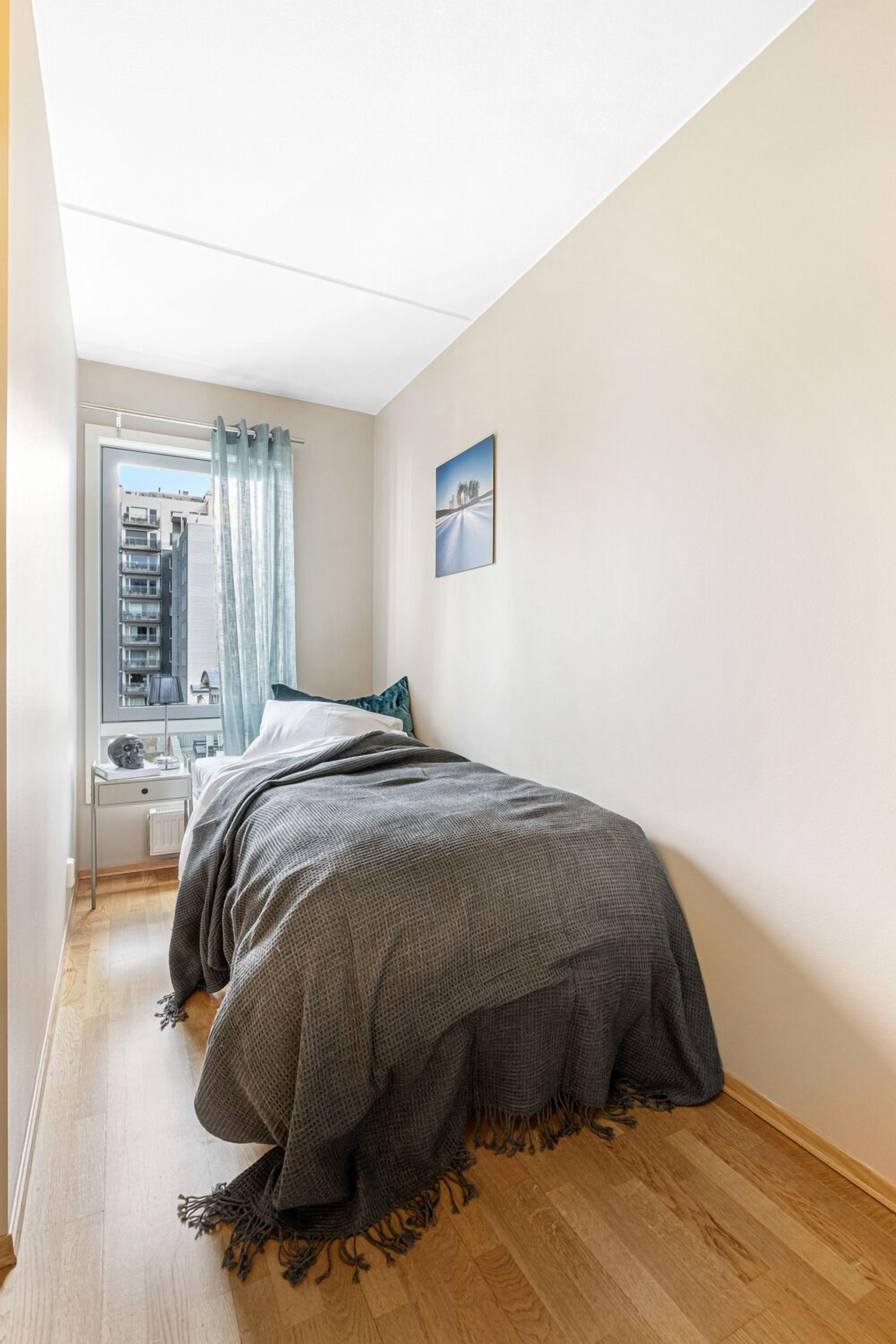 Una camera da letto con un letto, un comodino, una finestra con tende e un quadro appeso al muro, con vista sugli edifici circostanti.