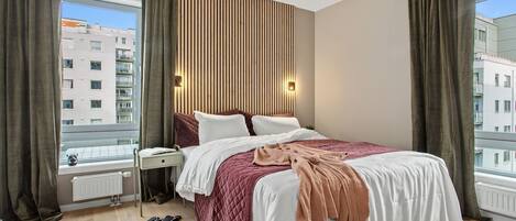 A modern hotel room with a large bed, a bedside table, a radiator, and a view of the city through large windows.