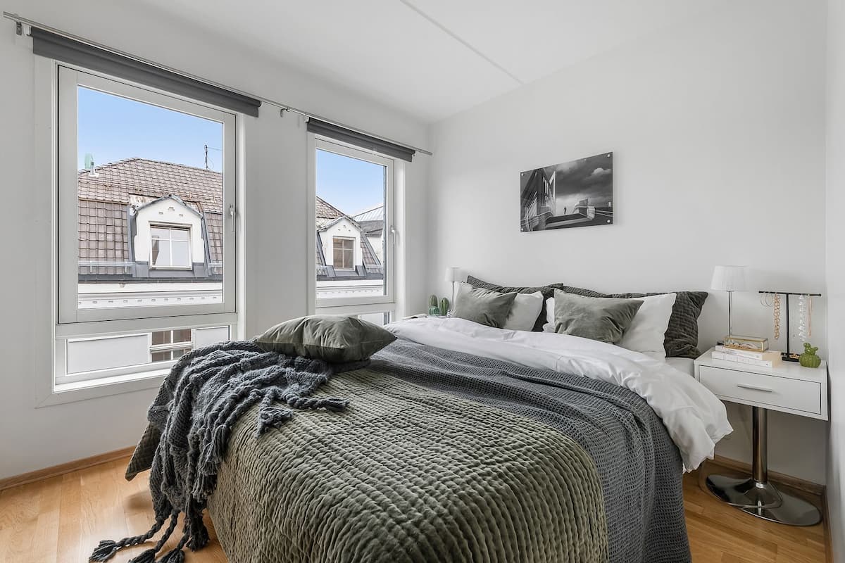 Une chambre à coucher avec un lit, des oreillers, une table de chevet, une lampe et une vue sur les bâtiments à travers de grandes fenêtres.