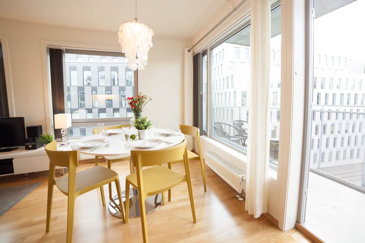 Un espace de salle à manger moderne avec une table ronde, des chaises jaunes, un lustre et une vue sur les bâtiments à travers de grandes fenêtres.