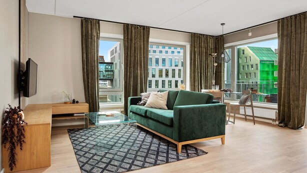 A modern living room with a green sofa, a glass coffee table, and a view of city buildings through large windows. - BJØRVIKA APARTMENTS, Teaterplassen, Oslo city center (Oslo)