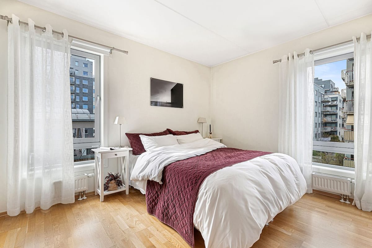 Une chambre avec un grand lit, deux tables de chevet, une fenêtre avec des rideaux et une vue sur des immeubles.