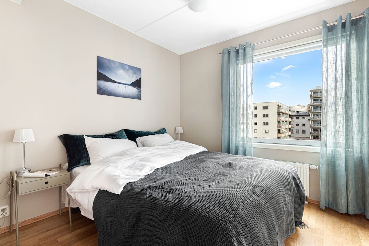 Une chambre moderne avec un grand lit, une table de chevet avec une lampe, une vue sur les immeubles par la fenêtre et un tableau accroché au mur.