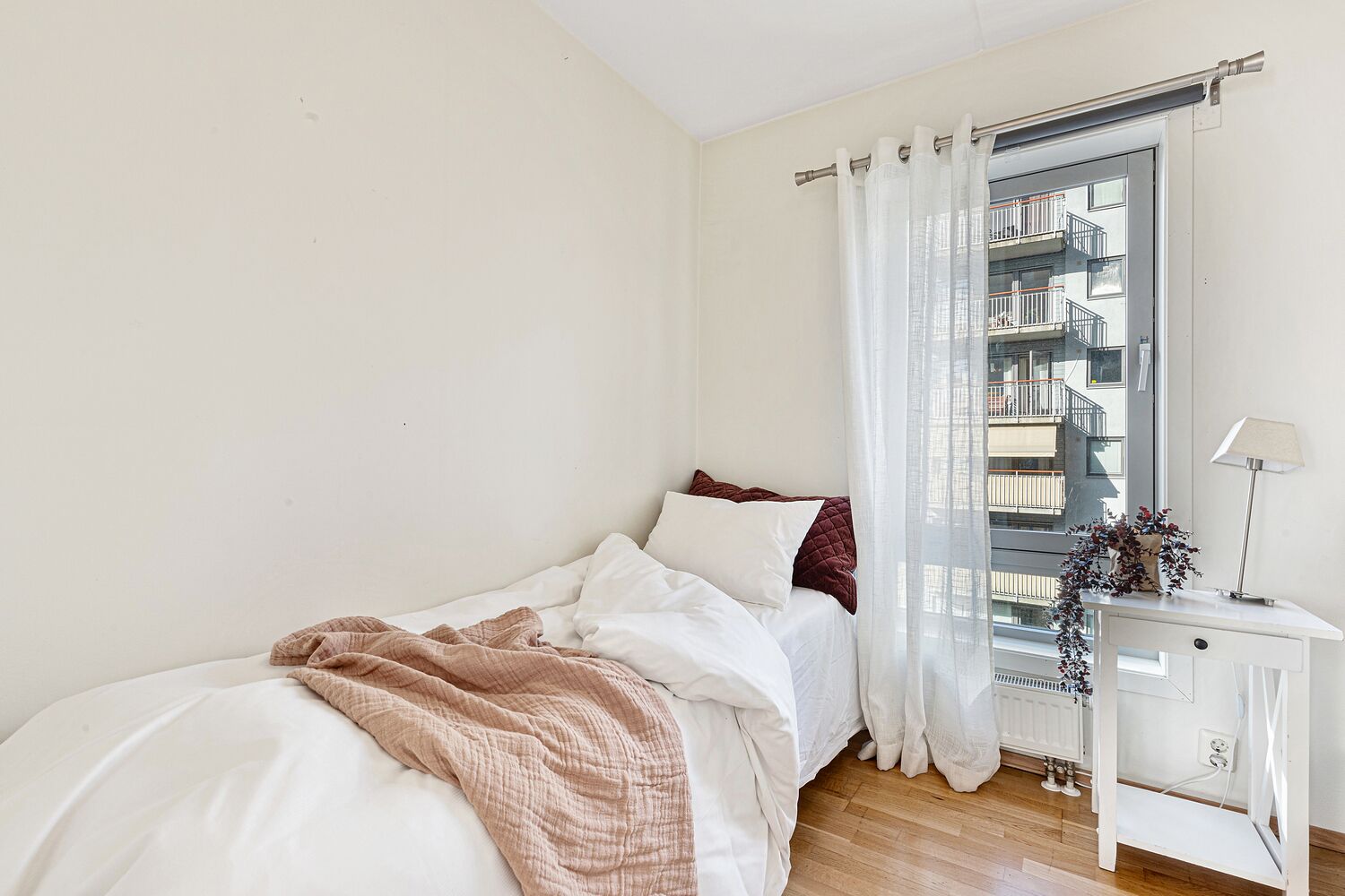 A bedroom with a bed, white curtains, a window with a view of buildings, and a bedside table with a lamp.