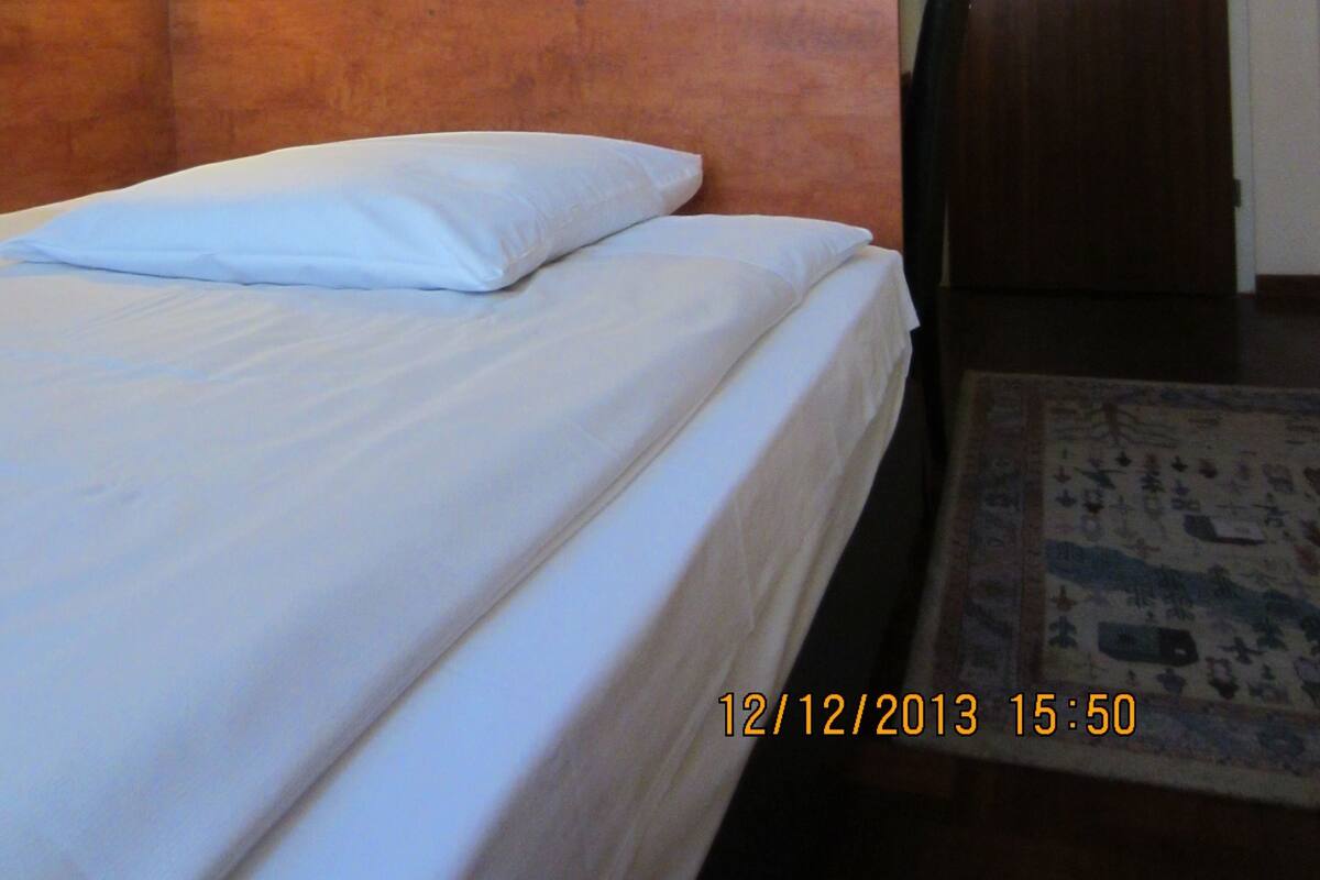 A hotel room with a bed, a nightstand, a wall-mounted lamp, a door, and a patterned rug.