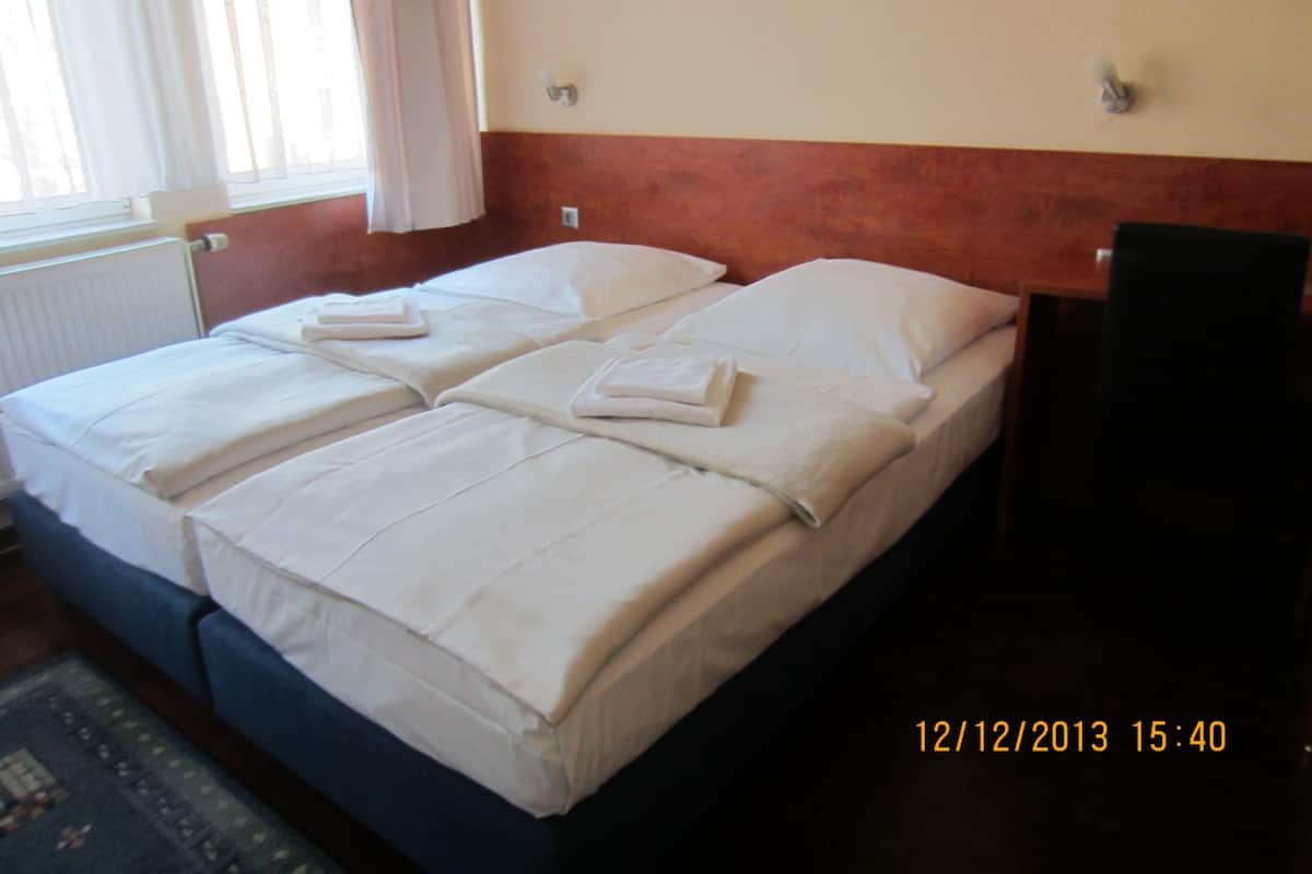 A double bed with white linens and pillows, a wooden headboard, a radiator, and a window with curtains.