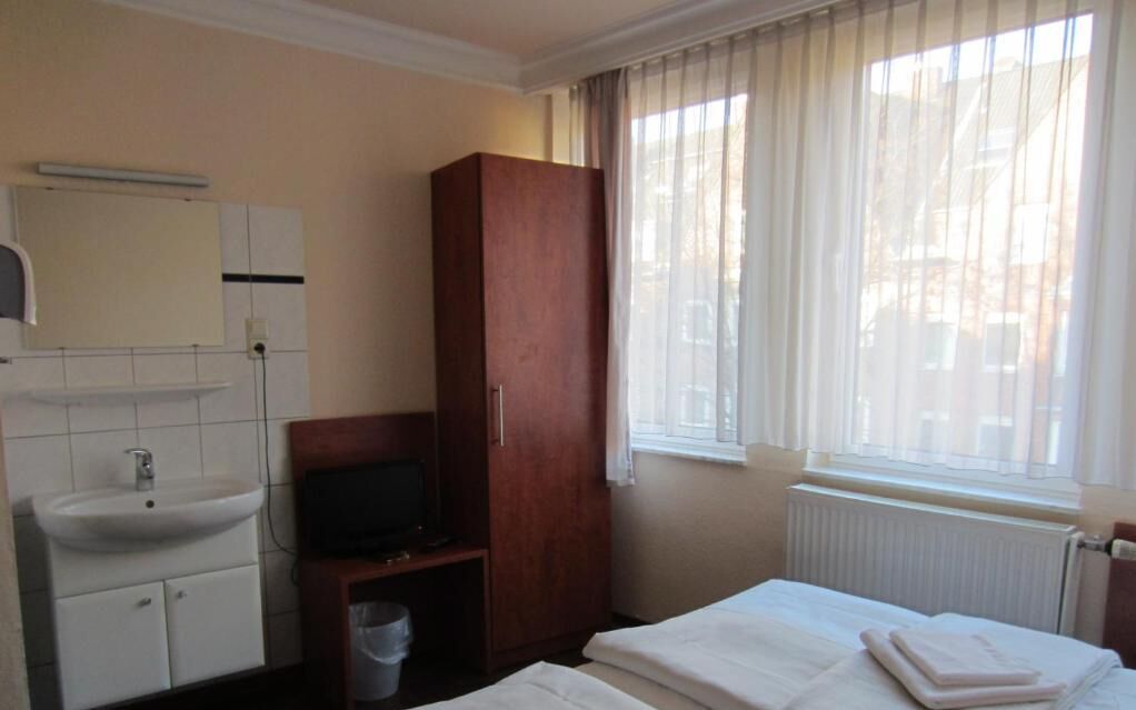 A hotel room with a bed, a wooden wardrobe, a television, a sink, and a window with curtains.