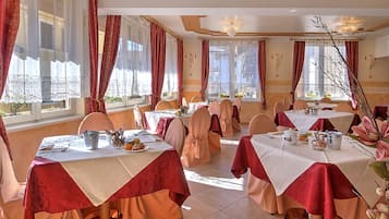 A dining area with tables set for service, large windows with curtains, and a chandelier.